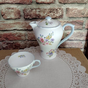 Può includere: Una teiera e una tazza da tè bianche con un motivo floreale in tonalità di rosa, blu e verde. La teiera ha un manico e un coperchio di colore azzurro chiaro. La tazza da tè ha un bordo e un manico di colore azzurro chiaro. Entrambi i pezzi sono su un sottopentola bianco.
