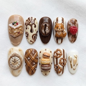 May include: A set of ten almond-shaped nail designs featuring various brown and cream-colored food-themed art. Designs include a stack of pancakes, chocolate swirls, a coffee bean, a fox, a raspberry, a clock, a pie crust, macarons, pretzels, and baking tools.