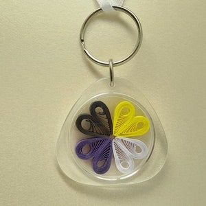 May include: A clear plastic keychain with a yellow, white, purple, and black paper flower inside. The flower is made of folded paper and is shaped like a circle with five petals.
