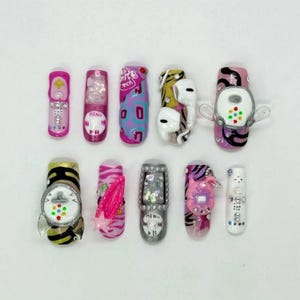 May include: A collection of twelve press-on nails with various designs. The nails feature a variety of colors, including pink, silver, and black, and are decorated with rhinestones, abstract patterns, and miniature electronic device designs.