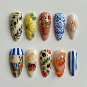 May include: A collection of ten artificial nails with various designs. The designs include olives, lemons, flowers in a pot, blue tile patterns, a seashell, a market scene, cheese and grapes, olives and tomatoes, a cocktail, and blue stripes.