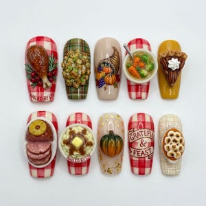May include: A set of ten Thanksgiving-themed nail art designs. Each nail features a different miniature food item, including a turkey leg, stuffing, a cornucopia, soup, pie, ham, mashed potatoes, a pumpkin, and a nail with the text "Grateful & Feast".