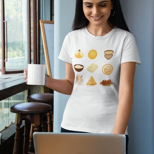 Op de afbeelding: Een wit T-shirt met een kleurrijke afbeelding van negen verschillende Indiase zoetigheden en desserts. De afbeelding bevat een kom kheer, een bord gulab jamun, een bord ladoo, een bord jalebi, een bord samosa, een bord rasgulla, een bord barfi, een bord halwa en een bord fruit.