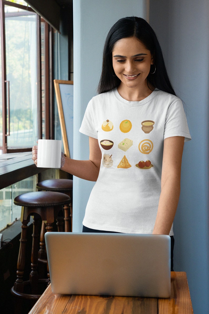 Op de afbeelding: Een wit T-shirt met een kleurrijke afbeelding van negen verschillende Indiase zoetigheden en desserts. De afbeelding bevat een kom kheer, een bord gulab jamun, een bord ladoo, een bord jalebi, een bord samosa, een bord rasgulla, een bord barfi, een bord halwa en een bord fruit.