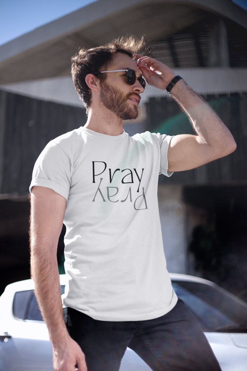 以下が含まれることがあります： 前面に黒で「Pray」という文字がプリントされた白いTシャツで、その下に「Pray」という文字が鏡文字でプリントされています。