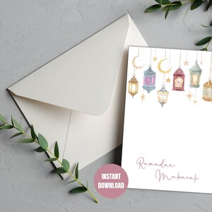 May include: Printable Ramadan Mubarak card with a white background and a string of colorful lanterns with gold stars and crescent moons. The text "Ramadan Mubarak" is written in pink cursive.