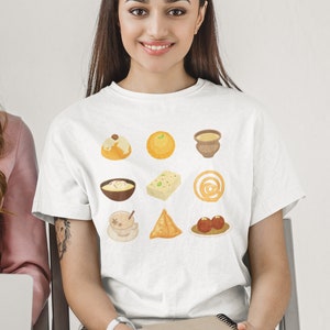 Op de afbeelding: Een wit T-shirt met een grafische print van negen verschillende Indiase zoetigheden en snacks. De afbeelding bevat een kopje chai, samosa's, jalebi, ladoo, gulab jamun en meer.