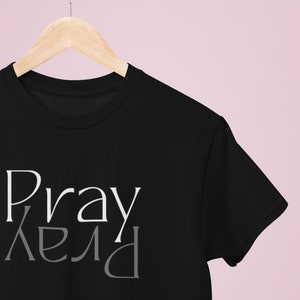 以下が含まれることがあります： 前面に白とグレーで「Pray」と書かれた黒いTシャツです。
