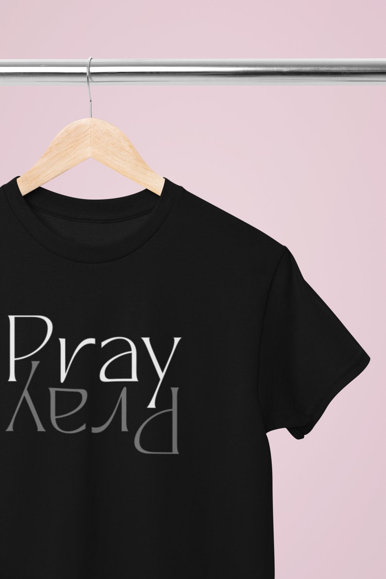 以下が含まれることがあります： 前面に白とグレーで「Pray」と書かれた黒いTシャツです。