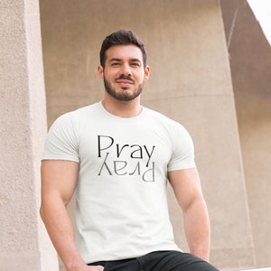 以下が含まれることがあります： 白いTシャツに黒い文字で「Pray」とグレーの文字で鏡面反射した「Pray」がプリントされています。