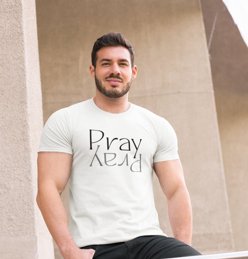 以下が含まれることがあります： 白いTシャツに黒い文字で「Pray」とグレーの文字で鏡面反射した「Pray」がプリントされています。