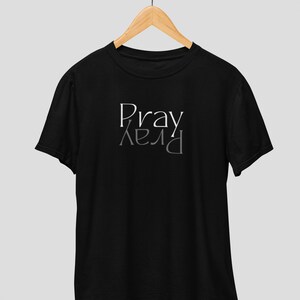 以下が含まれることがあります： 前面に白い文字で「Pray」とプリントされた黒いTシャツ。文字は鏡文字になっており、2回表示され、視覚効果を生み出しています。