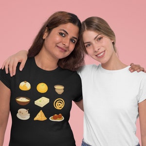 Op de afbeelding: Twee vrouwen dragen T-shirts. De vrouw links draagt een zwart T-shirt met een ontwerp van verschillende Indiase zoetigheden en snacks. De vrouw rechts draagt een wit T-shirt.