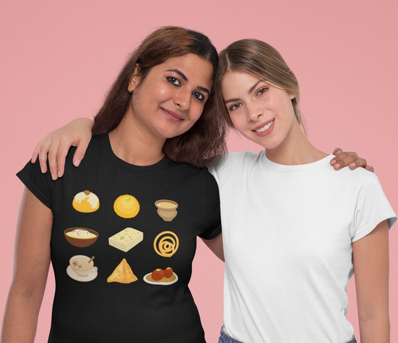 Op de afbeelding: Twee vrouwen dragen T-shirts. De vrouw links draagt een zwart T-shirt met een ontwerp van verschillende Indiase zoetigheden en snacks. De vrouw rechts draagt een wit T-shirt.