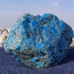 May include: A close-up of a vibrant blue mineral specimen. The rough, textured surface of the stone is a deep, saturated blue with hints of gray and black. The stone is displayed on a blue surface.