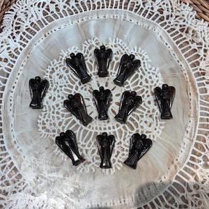May include: A collection of twelve dark brown angel figurines arranged in a circle on a white lace background.