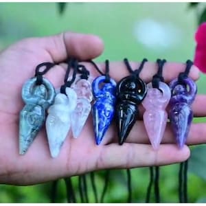 Könnte beinhalten: Eine Sammlung von sieben Anhängern aus geschnitztem Stein in Form stilisierter Figuren, die jeweils an einer schwarzen Kordel hängen. Die Anhänger haben verschiedene Farben, darunter Blau, Weiß, Transparent, Dunkelblau, Schwarz, Hellrosa und Violett.