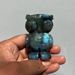 May include: A carved labradorite owl figurine featuring a dark grey base color with iridescent blue flashes across the surface. The owl has large circular eyes, a small beak, and defined wing details, showcasing the natural mineral texture.
