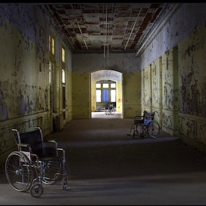 K&ouml;nnte beinhalten: Drei Rollst&uuml;hle stehen in einem langen, verlassenen Flur mit abbl&auml;tternder gelber Farbe und einer rissigen Decke. Der Flur ist schwach beleuchtet, und die Rollst&uuml;hle sind in unterschiedlichem Zustand der Vernachl&auml;ssigung.