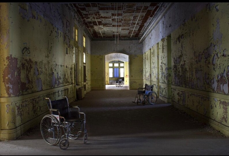 K&ouml;nnte beinhalten: Drei Rollst&uuml;hle stehen in einem langen, verlassenen Flur mit abbl&auml;tternder gelber Farbe und einer rissigen Decke. Der Flur ist schwach beleuchtet, und die Rollst&uuml;hle sind in unterschiedlichem Zustand der Vernachl&auml;ssigung.