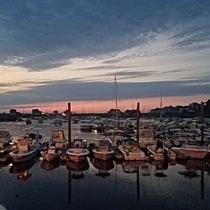 Może przedstawiać: Port pełen łodzi o zachodzie słońca. Niebo jest mieszanką różowego, pomarańczowego i niebieskiego. Łodzie są zacumowane przy molo i są różnych rozmiarów i kształtów.