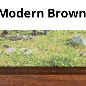 K&ouml;nnte beinhalten: Ein Nahaufnahme eines Holzbildrahmens mit brauner Oberfl&auml;che. Der Rahmen umgibt einen Druck eines gr&uuml;nen Grasfeldes mit Steinen.