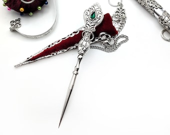 May include: A silver thimble with a green jewel on top, a red velvet pincushion, a silver needle threader with a chain, and a pair of silver scissors.