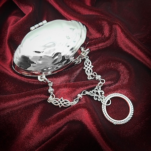 May include: A silver-colored, hammered-metal clutch with a decorative chain and ring. The clutch is oval-shaped with a hinged closure. The chain features Celtic knot designs and connects to a ring with a rope-like texture. The background is a deep red fabric.