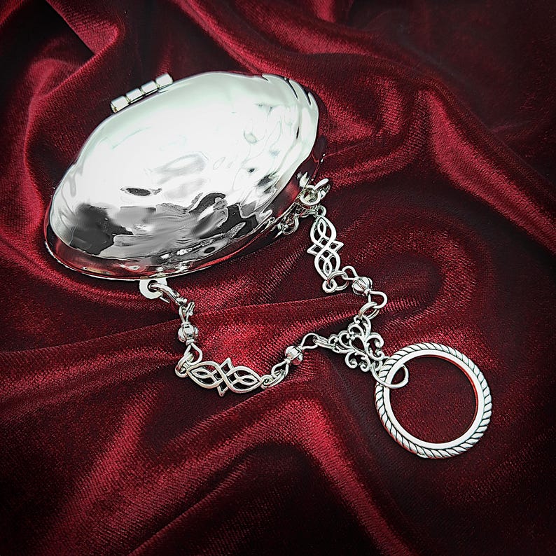 May include: A silver-colored, hammered-metal clutch with a decorative chain and ring. The clutch is oval-shaped with a hinged closure. The chain features Celtic knot designs and connects to a ring with a rope-like texture. The background is a deep red fabric.