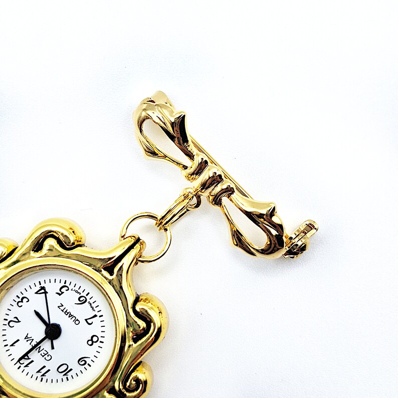 May include: Gold tone watch fob with a decorative bow pin and a round watch face with black hands and black Roman numeral markings. The watch face reads "GENEVA" and "QUARTZ".
