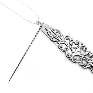 May include: A silver needle threader with a decorative, ornate design. The needle threader is open and a white thread is threaded through the loop.