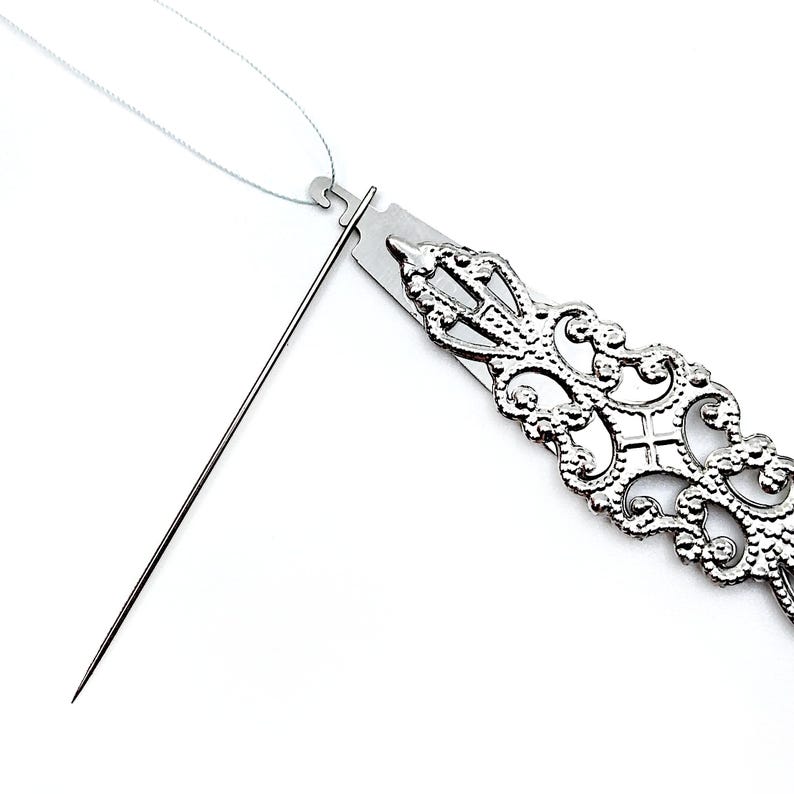 May include: A silver needle threader with a decorative, ornate design. The needle threader is open and a white thread is threaded through the loop.
