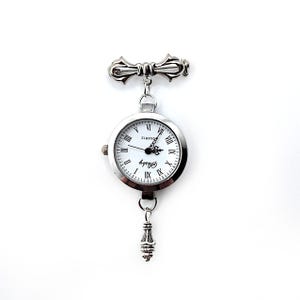 May include: Silver tone pocket watch with a white face and black Roman numeral hour markers. The watch is attached to a silver tone chain with a decorative clasp at the top.