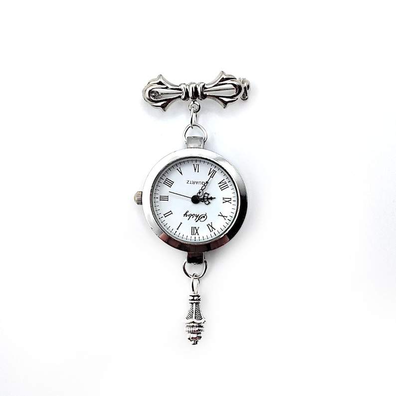 May include: Silver tone pocket watch with a white face and black Roman numeral hour markers. The watch is attached to a silver tone chain with a decorative clasp at the top.
