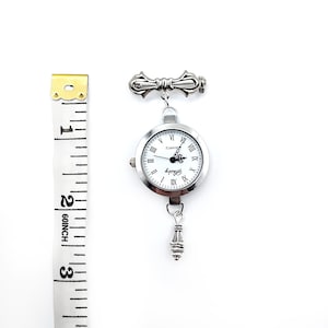 May include: A silver-toned watch with a white face and Roman numeral hour markers. The watch is attached to a silver-toned chain with a decorative pendant at the bottom. The watch is about 2 inches in diameter.