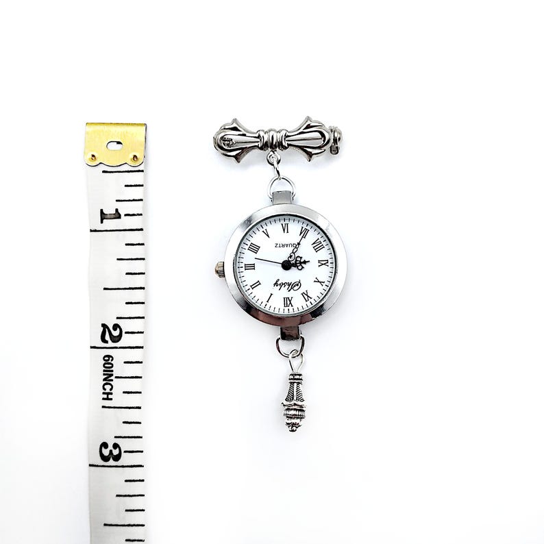 May include: A silver-toned watch with a white face and Roman numeral hour markers. The watch is attached to a silver-toned chain with a decorative pendant at the bottom. The watch is about 2 inches in diameter.