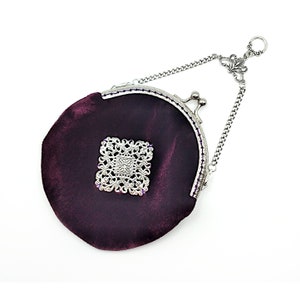 K&ouml;nnte beinhalten: Eine kleine, runde, violette Samt-Handtasche mit einer silbernen Kette und einem dekorativen silbernen Verschluss. Die Tasche hat ein silbernes Filigranmuster auf der Vorderseite.