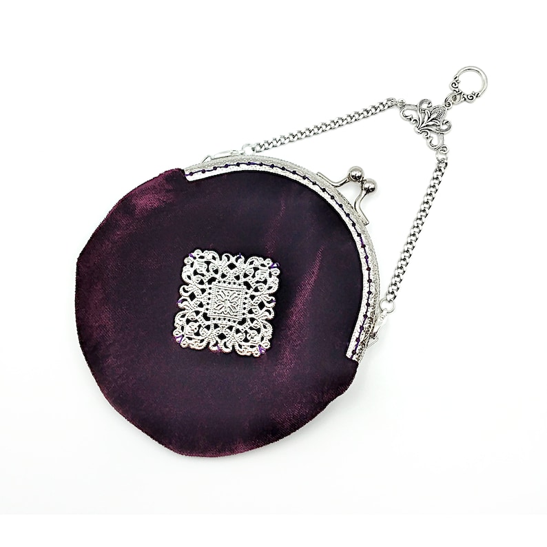 K&ouml;nnte beinhalten: Eine kleine, runde, violette Samt-Handtasche mit einer silbernen Kette und einem dekorativen silbernen Verschluss. Die Tasche hat ein silbernes Filigranmuster auf der Vorderseite.