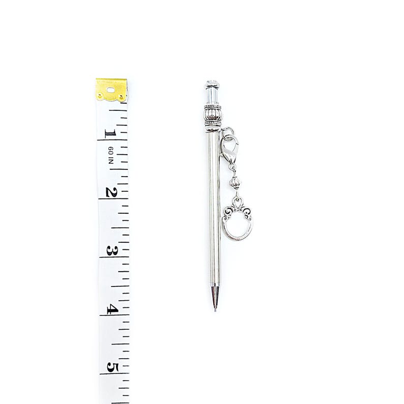 May include: A silver-toned mechanical pencil with a ribbed top section and a dangling charm attached by a lobster clasp. A white measuring tape is positioned alongside the pencil, indicating a length of approximately 4.5 inches.