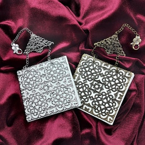 May include: Two small white journals with intricate silver and gold filigree designs on the covers. The journals are on a red velvet background.