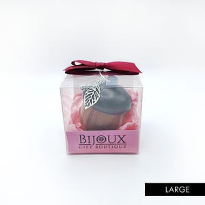 May include: A small, wooden acorn-shaped ornament with a silver leaf charm hanging from it. The ornament is packaged in a clear plastic box with a pink and red bow. The box has a pink label that reads "Bijoux Gift Boutique".