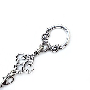 Pode incluir: Chaveiro prateado com detalhes ornamentais em volutas. O chaveiro apresenta um formato de chave decorativa e um fecho circular. O design &eacute; definido em um fundo branco brilhante, destacando o trabalho em metal.