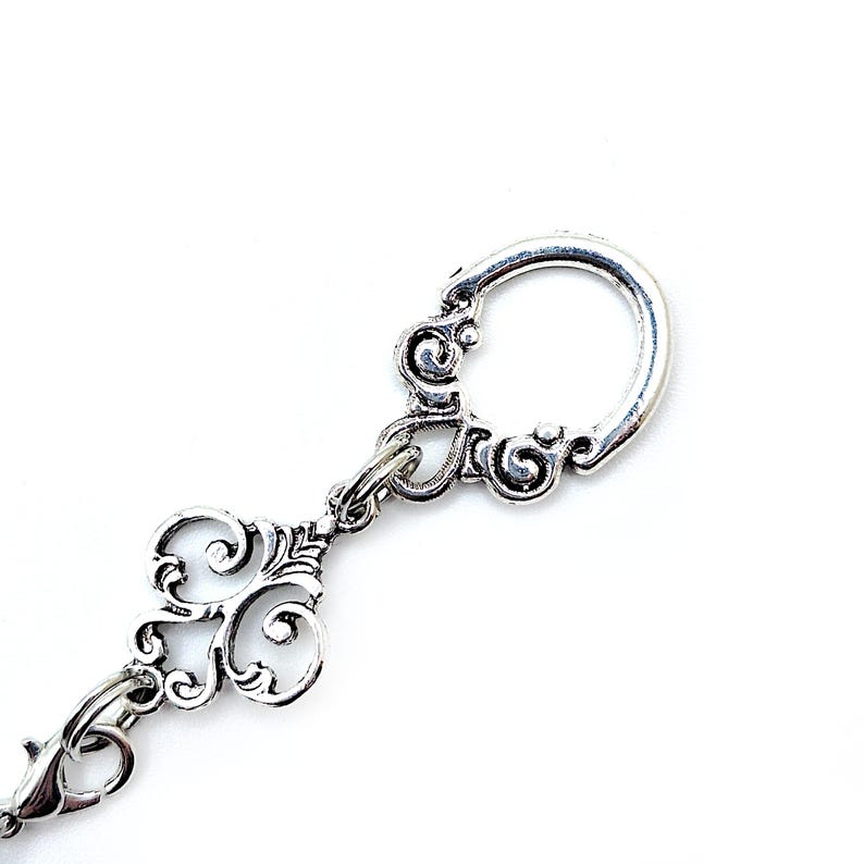Pode incluir: Chaveiro prateado com detalhes ornamentais em volutas. O chaveiro apresenta um formato de chave decorativa e um fecho circular. O design &eacute; definido em um fundo branco brilhante, destacando o trabalho em metal.