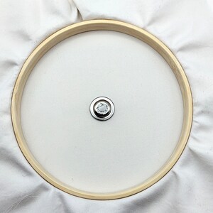 May include: A white fabric stretched over a wooden embroidery hoop with a metal screw in the center.