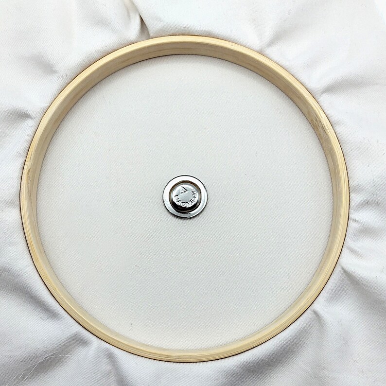 May include: A white fabric stretched over a wooden embroidery hoop with a metal screw in the center.