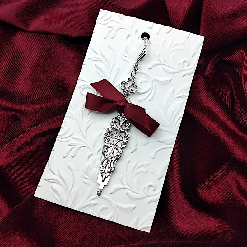 May include: A silver bookmark with a decorative design and a burgundy ribbon bow. The bookmark is on a white card with a floral pattern.