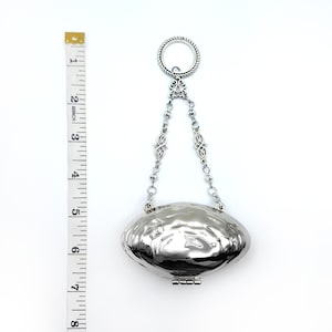 May include: A silver-colored, oval-shaped pendant with a textured surface and a hinged bottom. The pendant is suspended from a chain with decorative links and a ring at the top. The pendant is approximately 3 inches long.