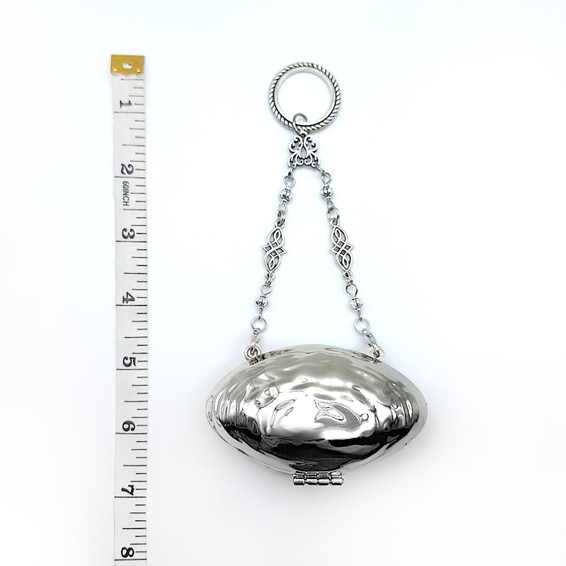 May include: A silver-colored, oval-shaped pendant with a textured surface and a hinged bottom. The pendant is suspended from a chain with decorative links and a ring at the top. The pendant is approximately 3 inches long.