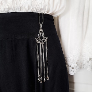 May include: A silver-toned skirt clip with a decorative floral design and dangling chains. The clip is attached to a black skirt, with a white blouse visible in the background. The chains end with small, decorative charms.