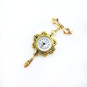 May include: Gold-toned pocket watch with a white face and black hands. The watch is attached to a gold-toned chain with a decorative key charm.
